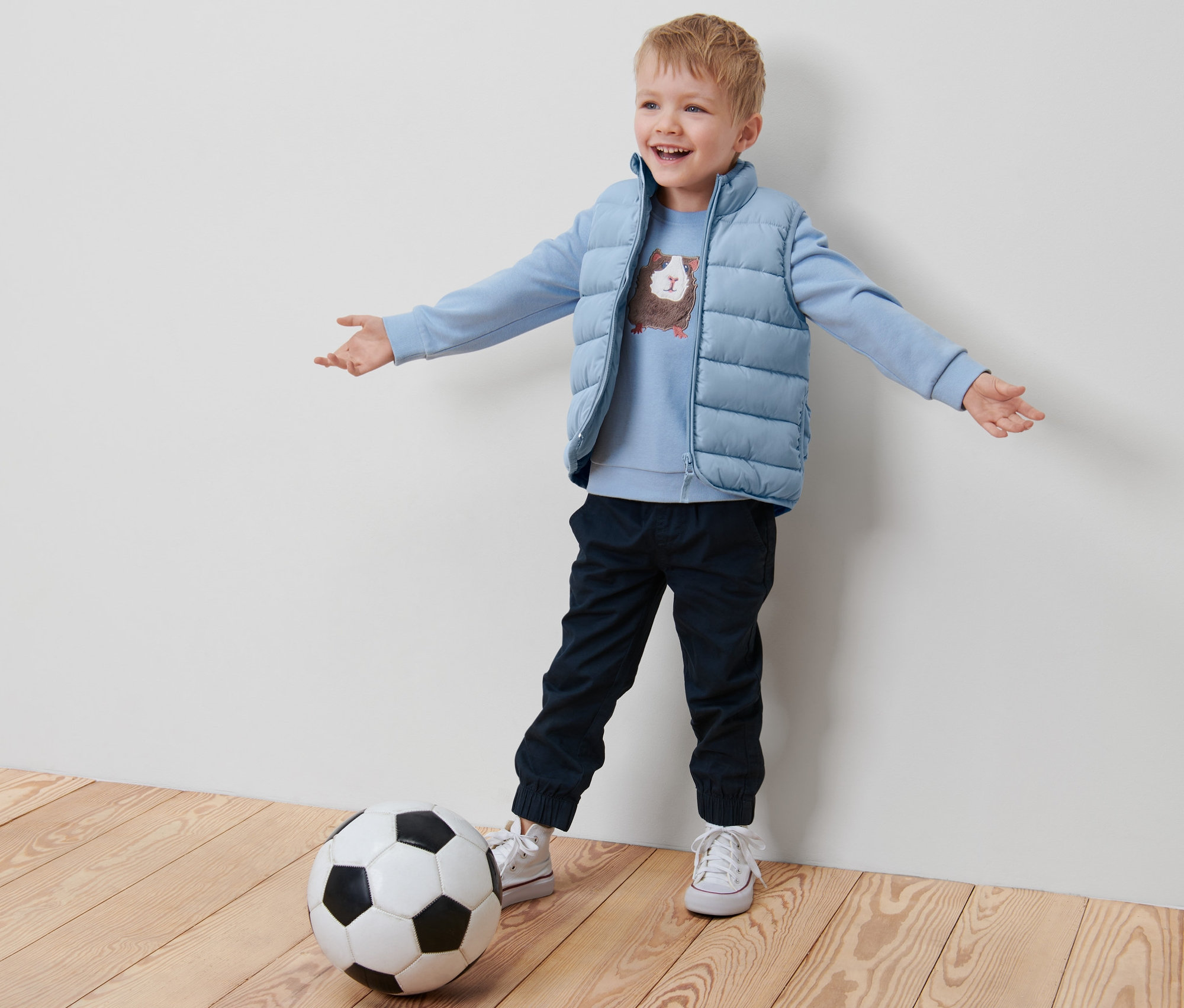 Ein Junge steht mit ausgestreckten Armen neben einem Fußball, er trägt eine blaue Kleinkind-Steppweste.