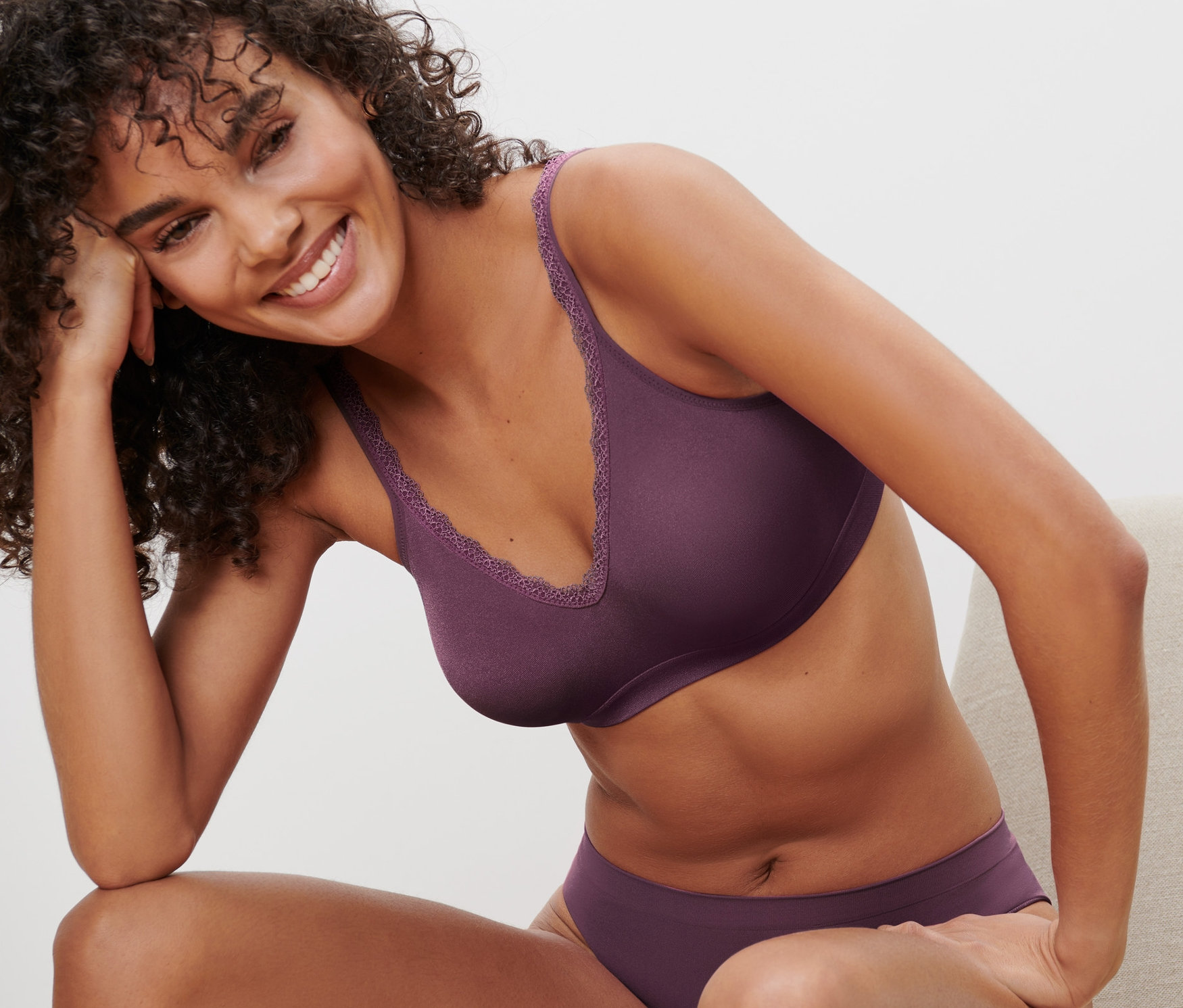 Frau sitzt in auberginefarbenem Seamless-Soft-BH und passendem Höschen auf einem Stuhl.