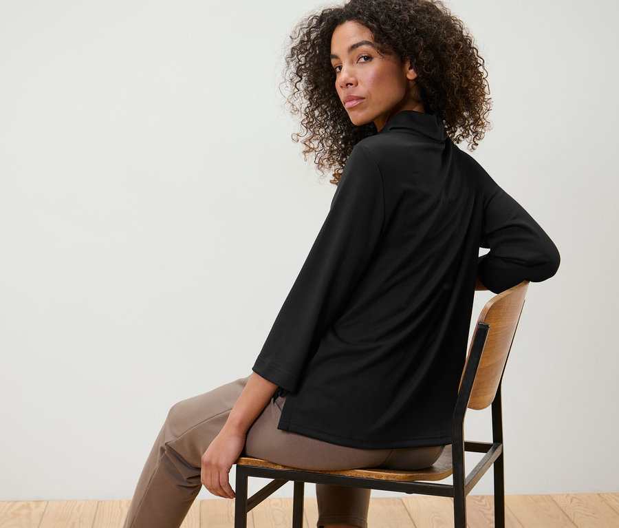 Frau sitzt auf einem Stuhl und trägt ein schwarzes Sweatshirt mit 3/4-Arm.