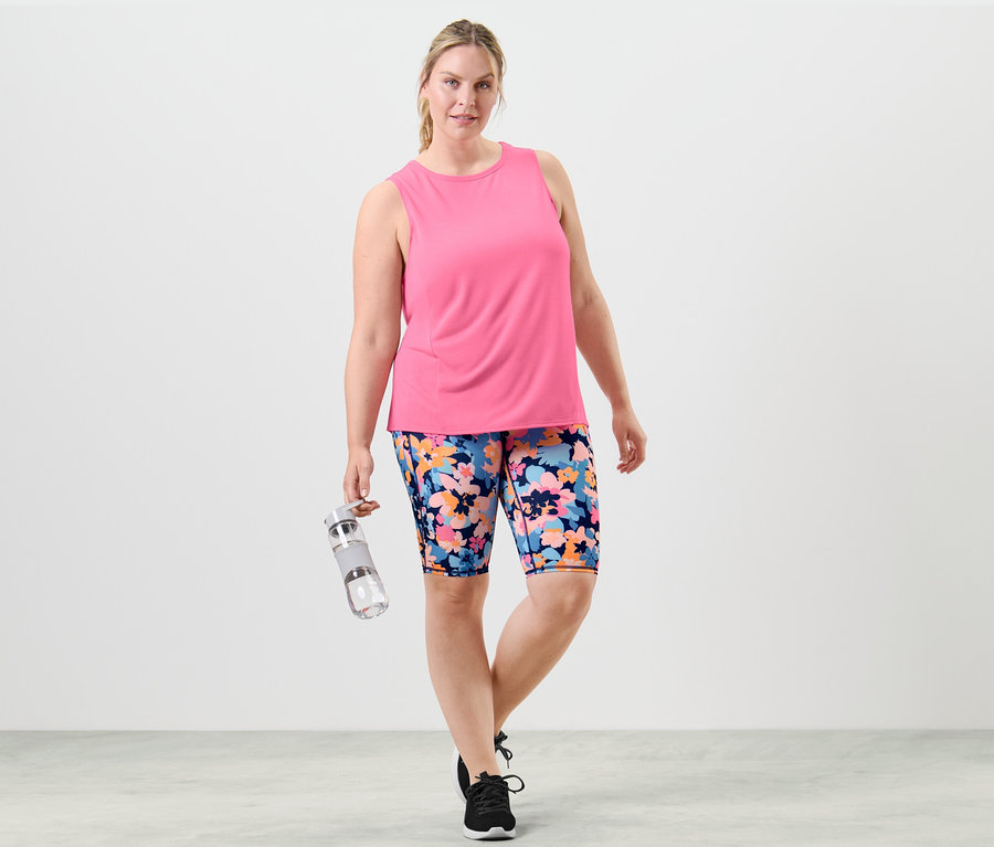 Frau posiert in Sportkleidung mit einem pfirsichfarbenen Top und geblümten Shorts und hält eine Wasserflasche.