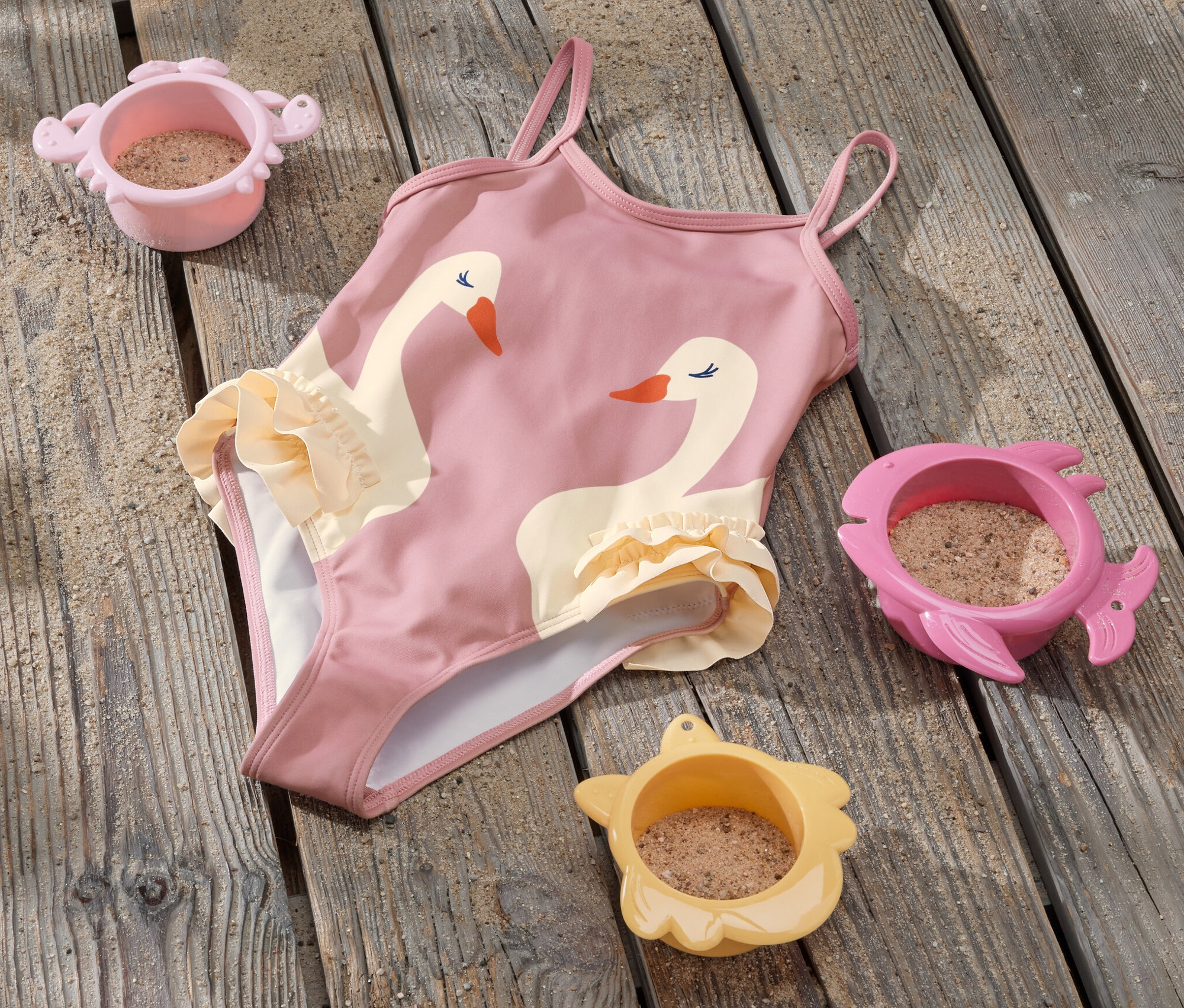 Ein Kinder-Badeanzug mit Rüschen und Schwanenmotiv liegt auf einem Holzboden neben drei Sandspielzeugen.