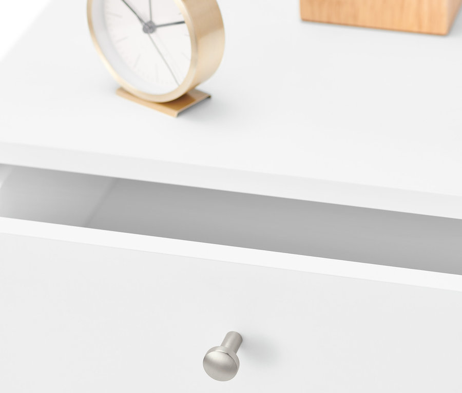 Detailansicht eines weißen Nachttisches mit offener Schublade, goldener Uhr und einer Holzkiste.