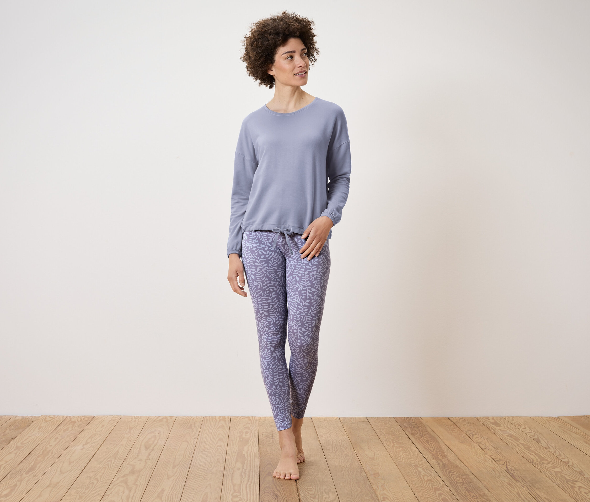 Frau in hellblauem Schlafanzug mit Blattmuster steht auf Holzboden.