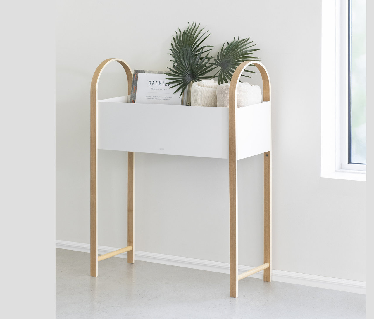 Umbra Bellwood Blumenkasten und Aufbewahrungsbox steht neben einem Fenster an einer Wand, darin sind Pflanzen und Handtücher.