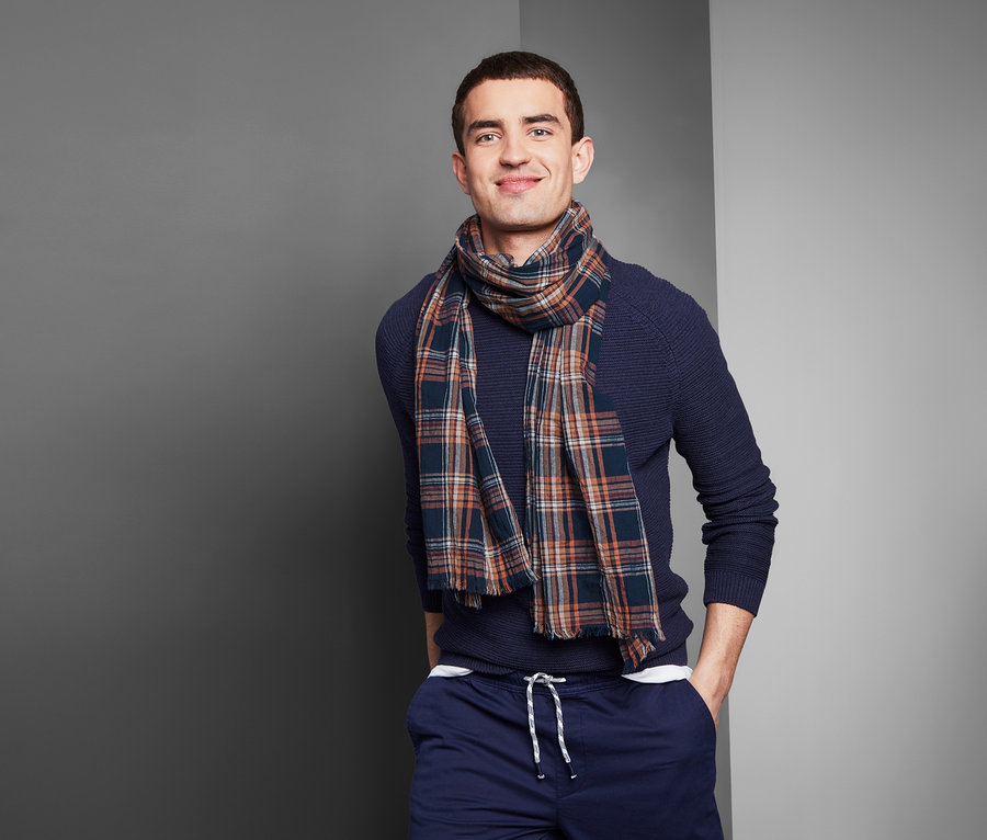Lächelnder Mann posiert in blauem Pullover und kariertem Webschal.