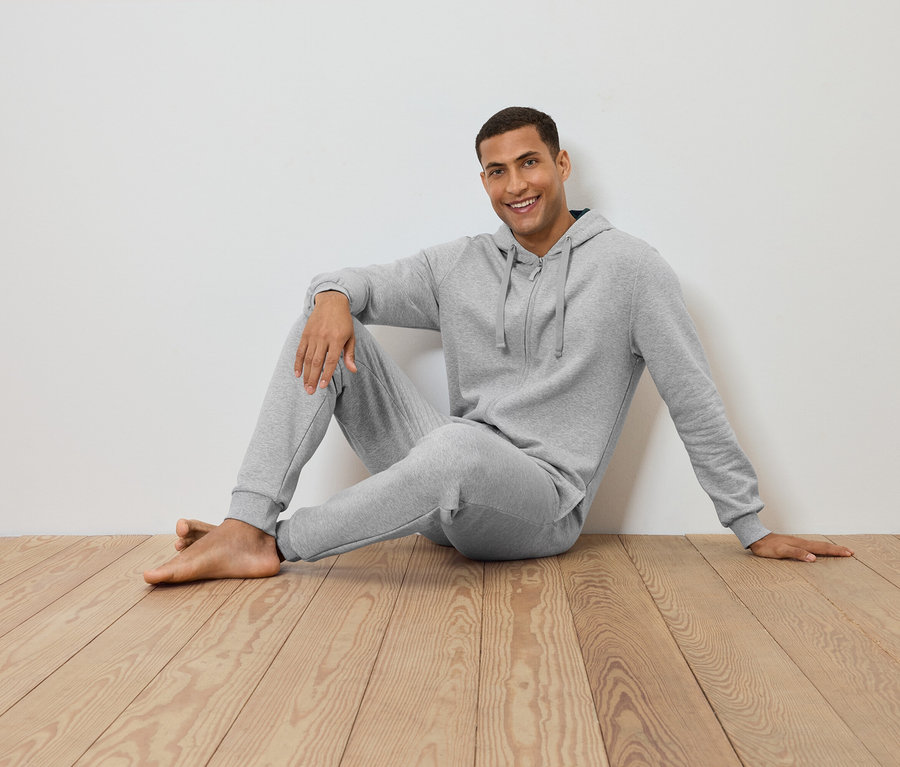 Mann sitzt auf Holzboden an einer weißen Wand, trägt einen grauen Hoodie und eine Jogginghose.