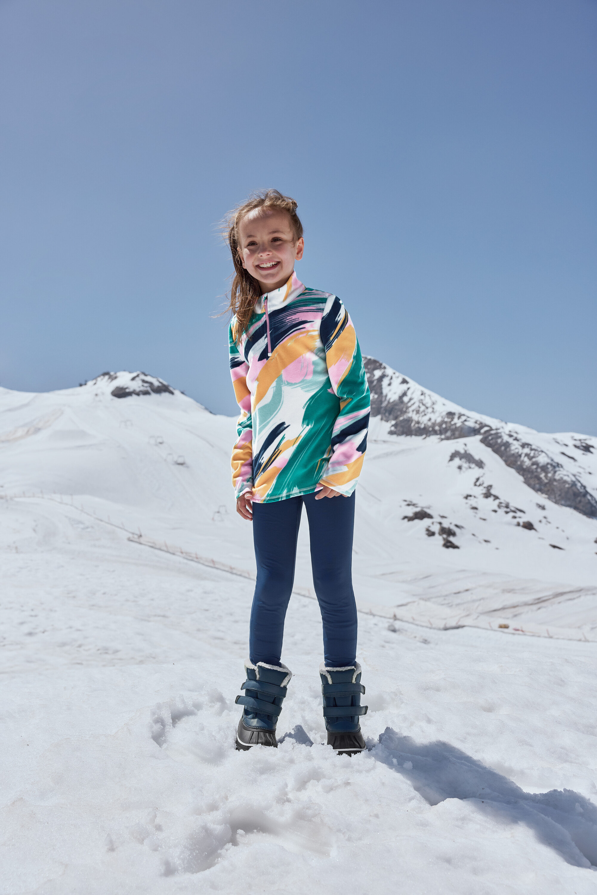 Ein lächelndes Mädchen steht im Schnee und trägt blaue Kinder-Allwetterstiefel.