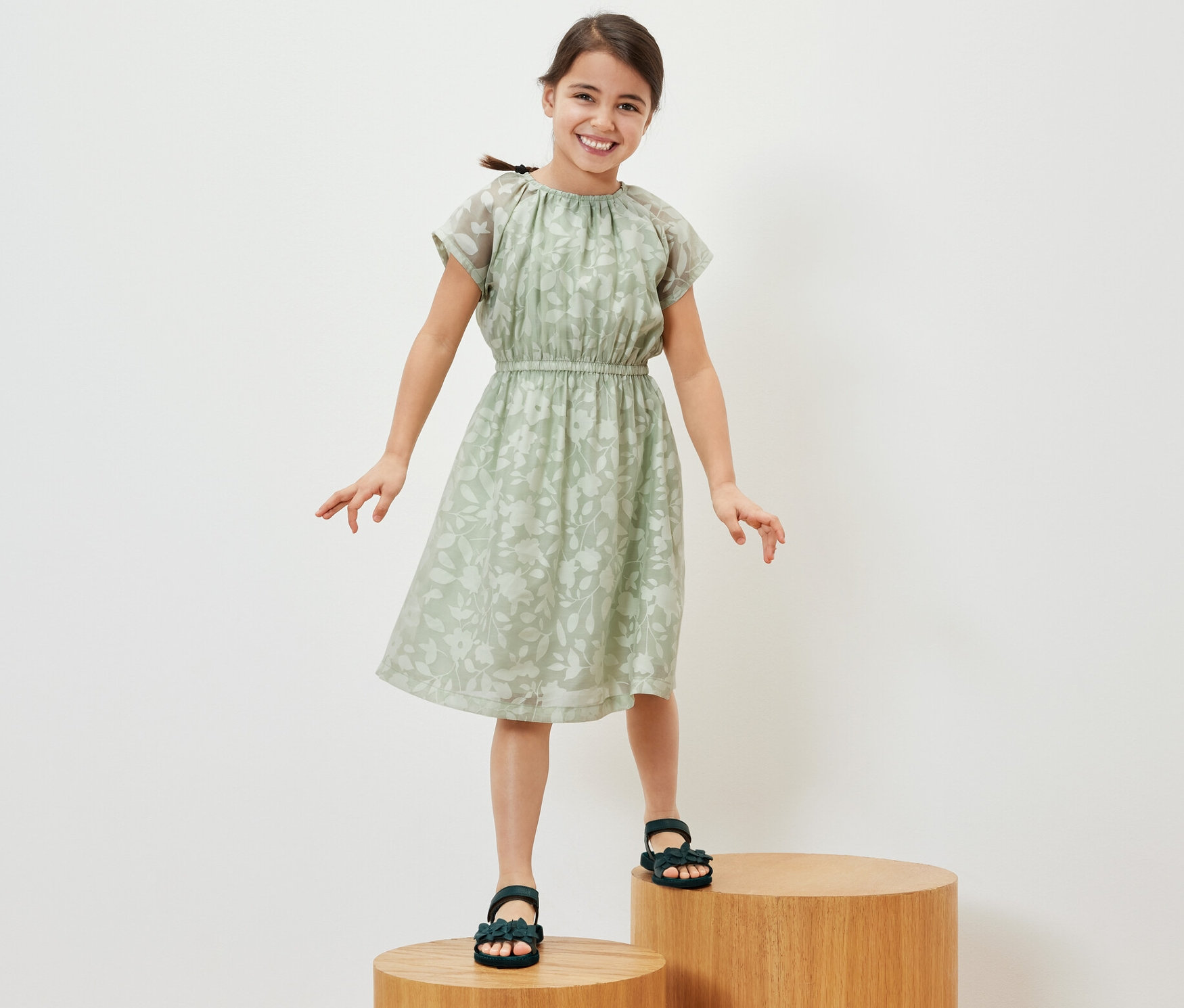 Ein lächelndes Mädchen steht auf Holzstufen und trägt ein Kinder-Kleid.