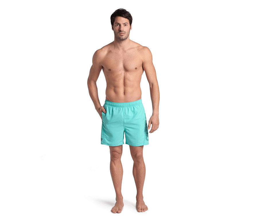 Mann steht mit der Hand in der Tasche der hellblauen Badeshorts arena Fundamentals Logo.