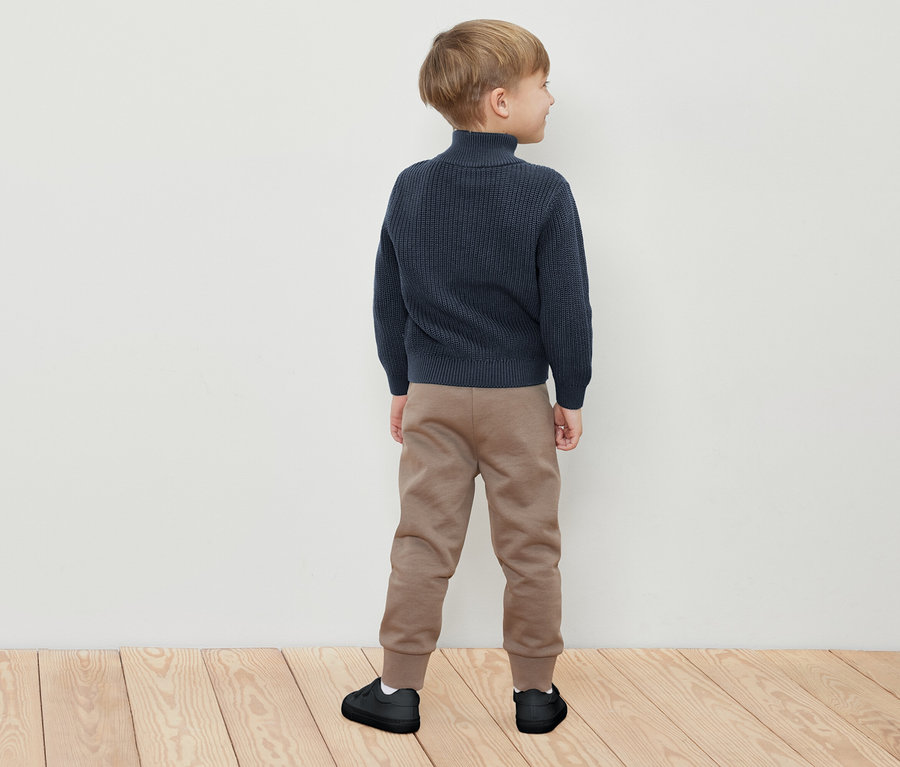 Ein Junge in einem dunkelblauen Pullover, beigen Jogginghosen und schwarzen Schuhen steht mit dem Rücken zum Betrachter.