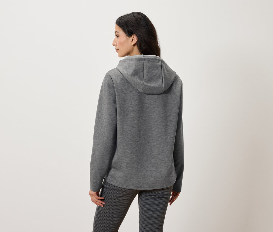 Rückansicht einer Frau mit grauem Kapuzenpullover und gemusterter Hose.