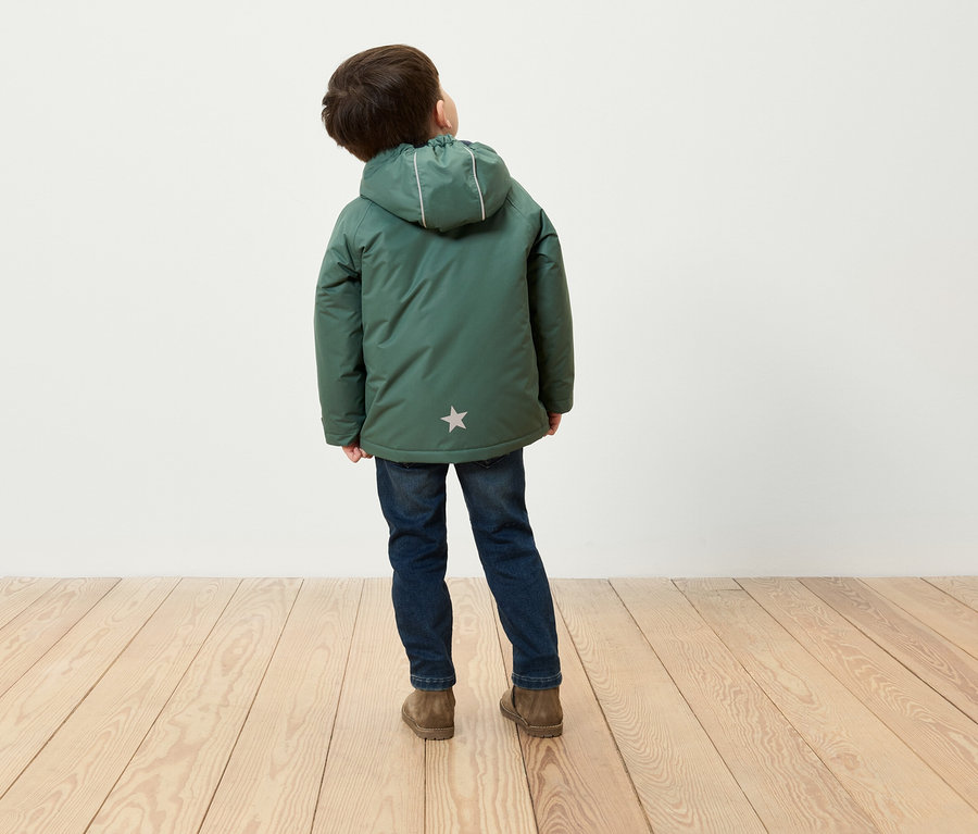 Junge mit dem Rücken zur Kamera trägt eine grüne Winterjacke mit Kapuze, blaue Jeans und braune Schuhe und steht auf einem Holzboden.