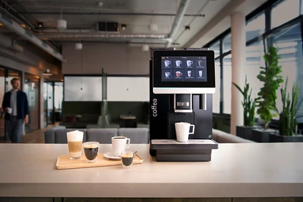 Vier Tassen Kaffee auf einem Holzbrett vor einer Kaffeemaschine in einem Büro.