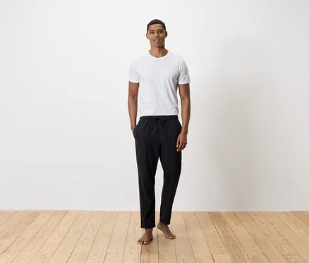 Ein Mann steht barfuß auf einem Holzboden und trägt ein weißes T-Shirt und eine schwarze Hose.