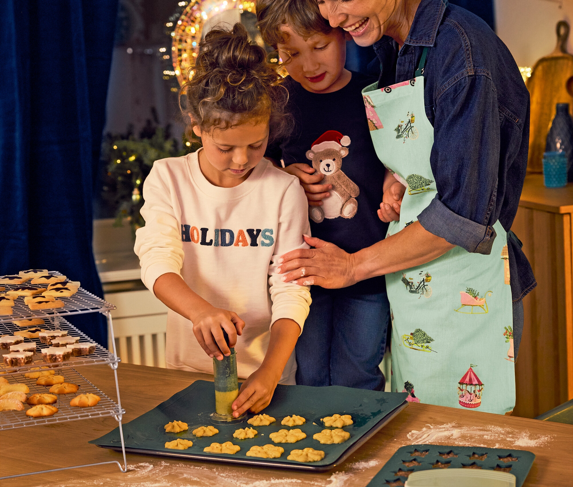 Eine Mutter backt mit ihren Kindern Kekse. Ein Mädchen im Baumwoll-Sweatshirt sticht Formen aus dem Teig.