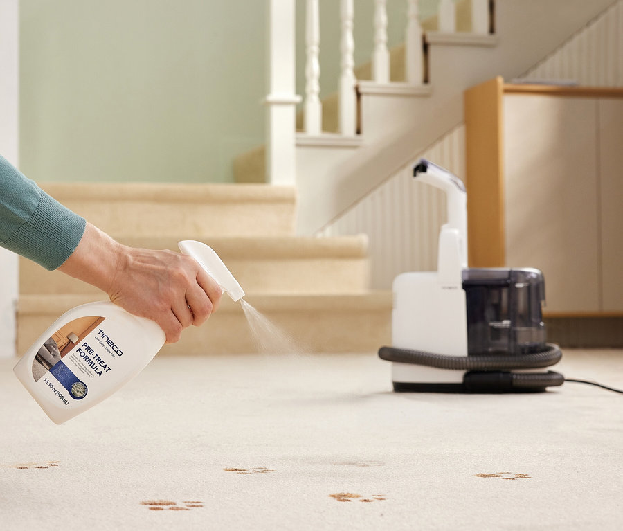 Eine Frau sprüht Tineco i Carpet Spot auf Flecken auf einem Teppich, im Hintergrund eine Treppe.