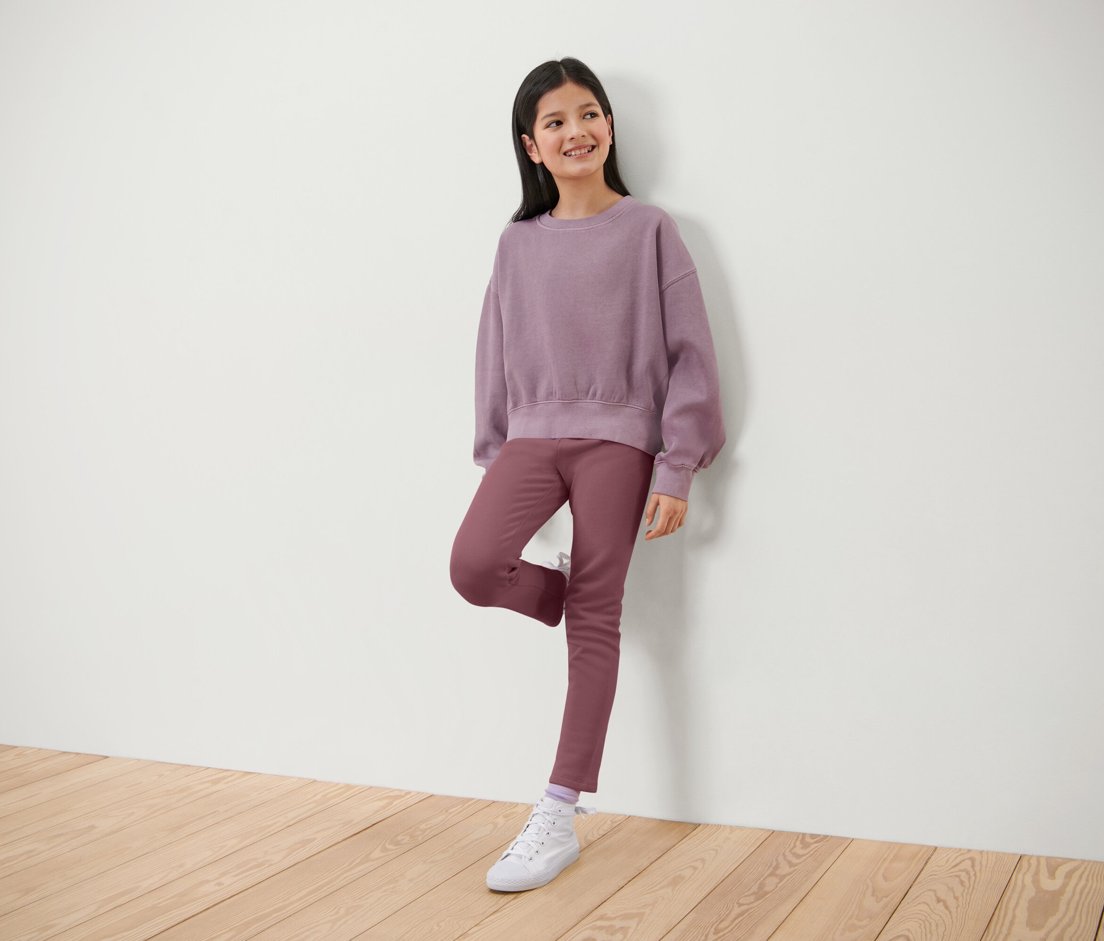 Lächelndes Mädchen posiert in lila Pullover und Hose, lehnt an einer weißen Wand.