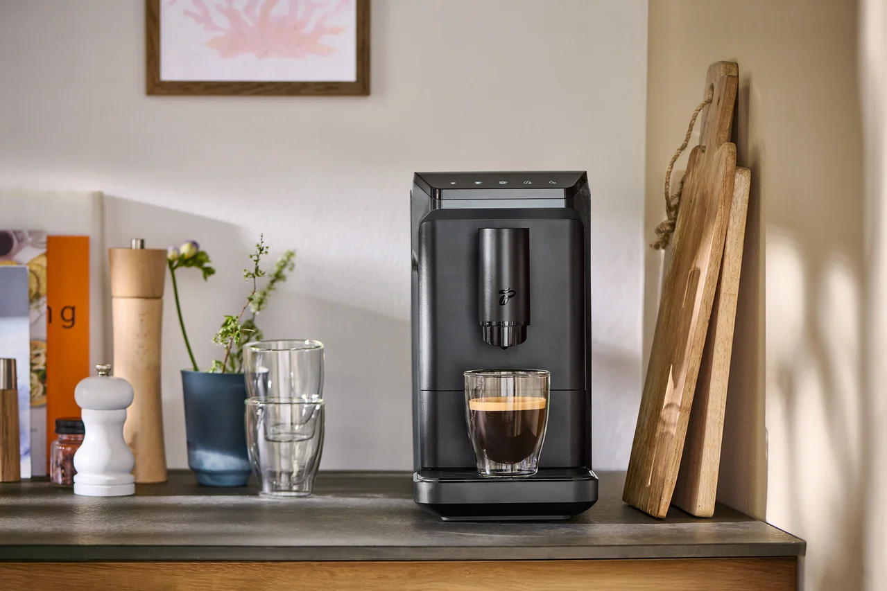 Grauer »Esperto mini« Kaffeevollautomat füllt Kaffee in ein Glas, daneben Schneidebretter, Gewürzmühlen und Gläser auf einer Arbeitsplatte.