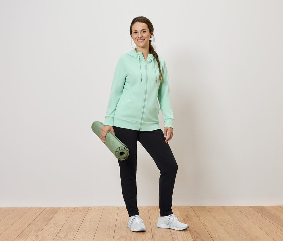 Frau hält eine zusammengerollte grüne Yogamatte, trägt einen mintfarbenen Kapuzenpullover und eine schwarze Trainingshose.