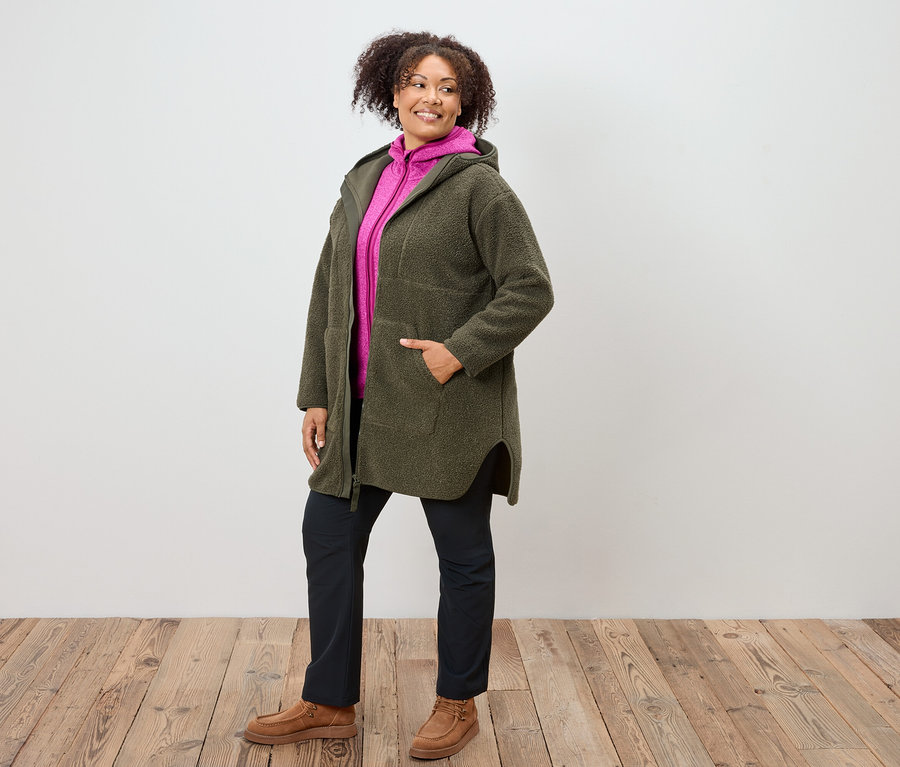 Frau in olivgrünem Kapuzenmantel, pinkfarbenem Hoodie, marineblauer Hose und braunen Schuhen steht auf Holzboden.