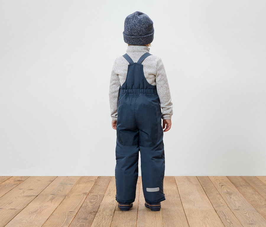 Junge mit dem Rücken zur Kamera trägt blaue Latzhose, grauen Pullover und blaue Mütze.