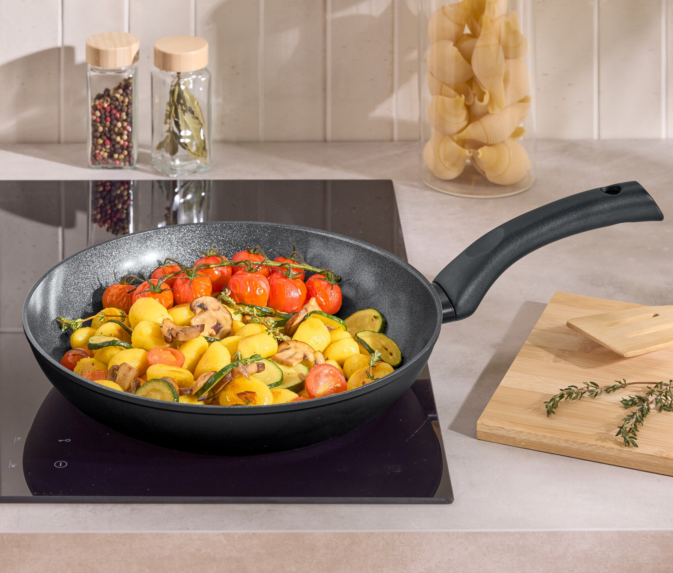 In einer Bratpfanne auf einem Kochfeld werden Kartoffeln, Zucchini, Champignons und Cherrytomaten gebraten. Daneben liegen ein Holzbrett und ein Pfannenwender.