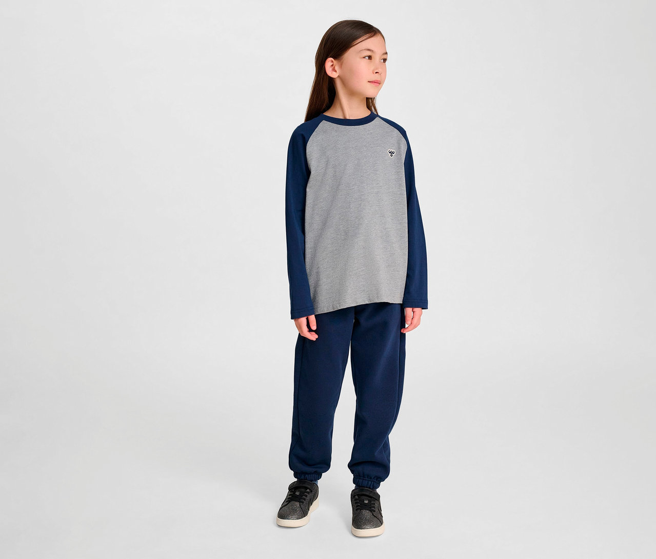 Mädchen steht und trägt eine blaue Trainingshose und ein graues HUMMEL HMLJR Raglan Loose T-Shirt BEE.