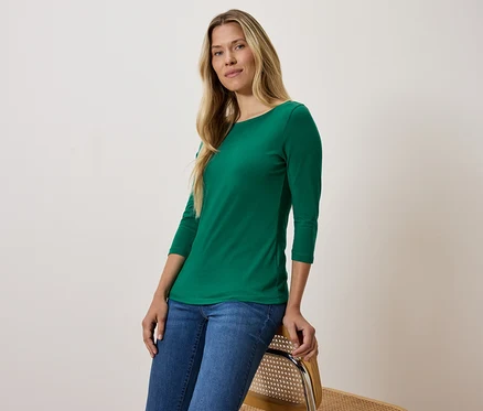 Blonde Frau in grünem Dreiviertelarm-Shirt und blauen Jeans sitzt auf einem Stuhl.