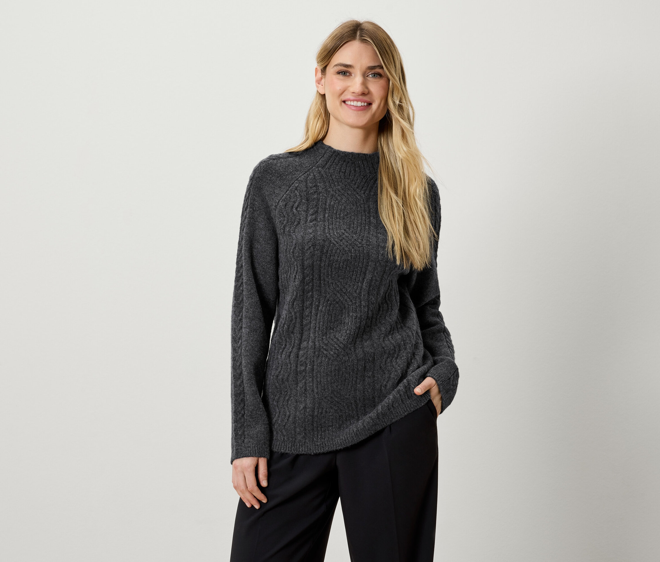 Eine Frau posiert in einem grauen Strickpullover und einer schwarzen Hose.