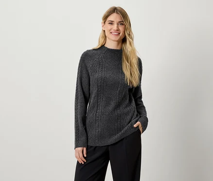 Eine Frau posiert in einem grauen Strickpullover und einer schwarzen Hose.