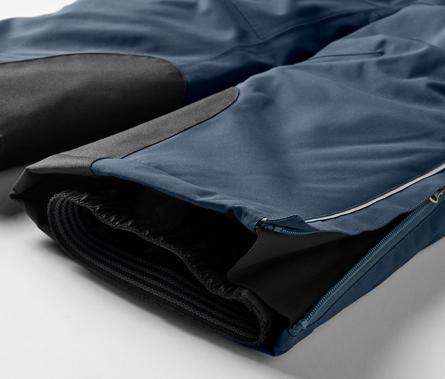 Detail einer blauen Hose mit schwarzen Knieeinsätzen und einem Reißverschluss am Bein.