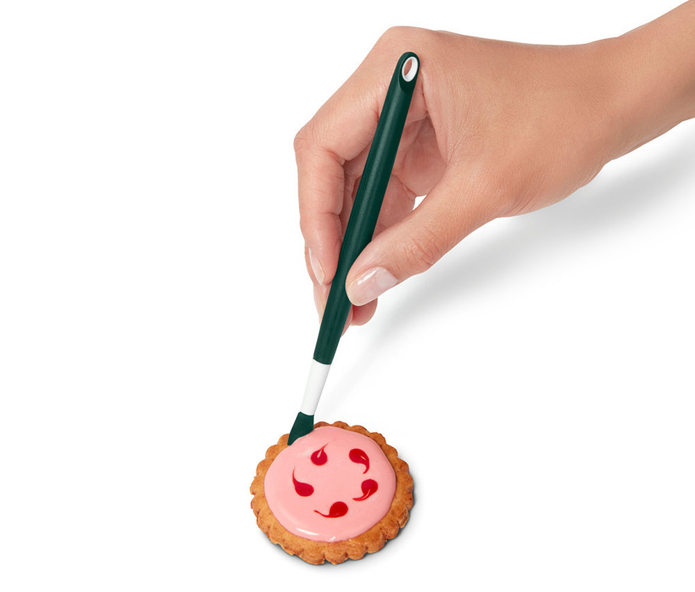 Eine Hand verziert einen Keks mit rosa Glasur mit einem grünen Dekorstift.