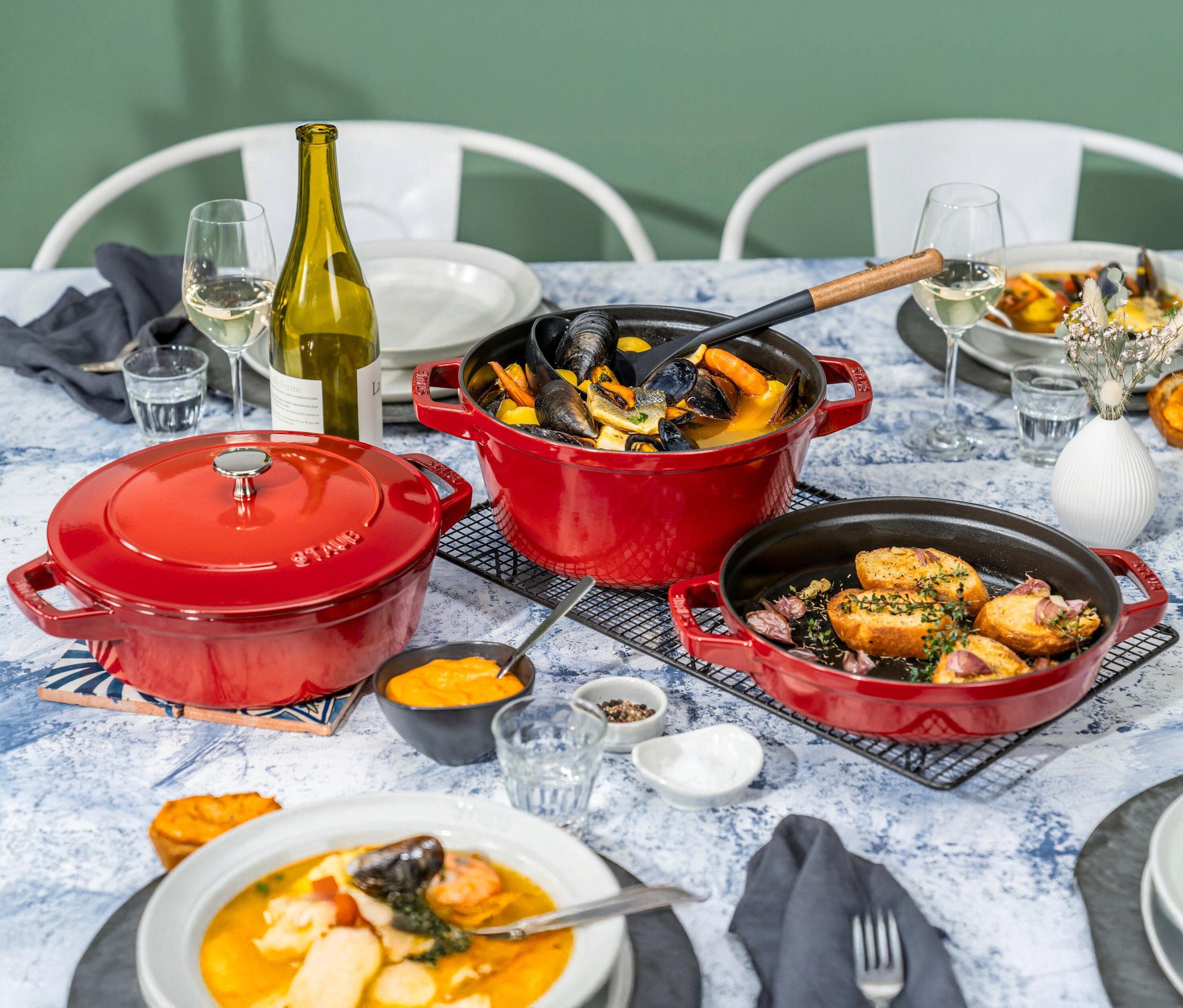 Gedeckter Tisch mit Speisen in drei roten STAUB Kochtöpfen »La Cocotte«, 3-teilig, einer Weinflasche und Tellern.