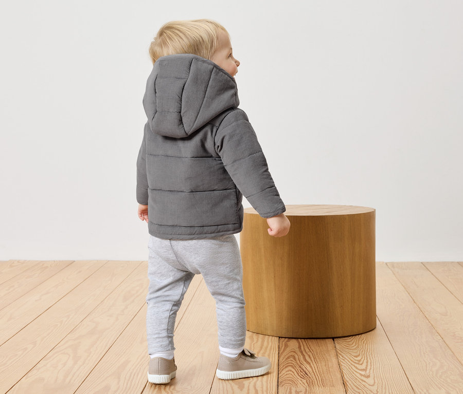Kleiner Junge mit blonden Haaren trägt eine graue, gesteppte Jacke mit Kapuze, graue Hose und beigefarbene Schuhe. Er steht auf einem Holzboden neben einem runden Holzhocker.