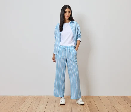 Frau in blau-weiß gestreifter Hose und Hemd über einem weißen T-Shirt, mit weißen Turnschuhen auf Holzboden vor einer weißen Wand.