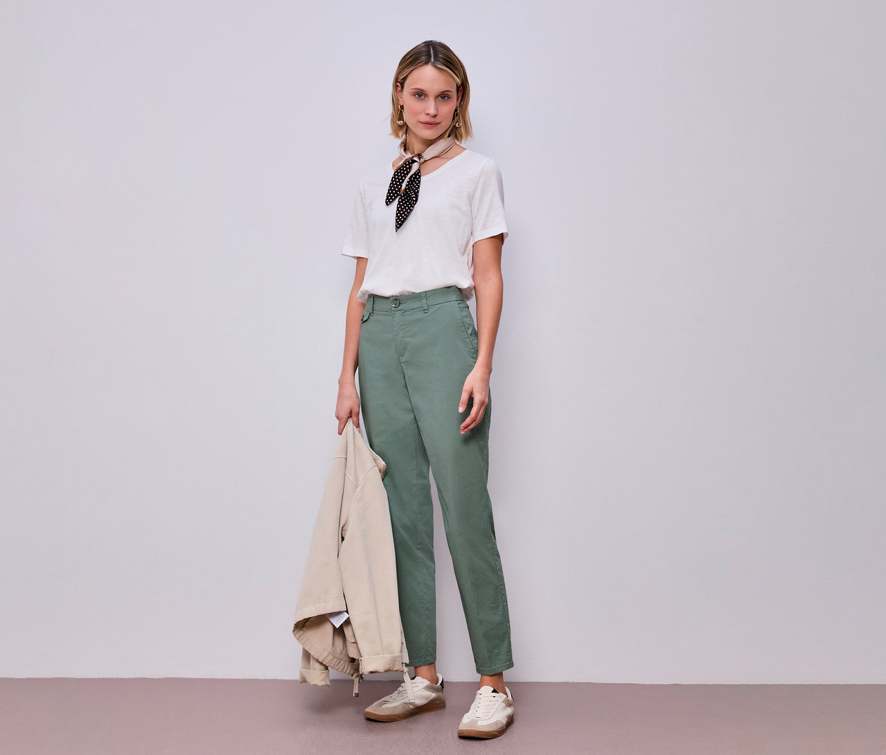 Frau in mintfarbener Street One Chino mit T-Shirt und Halstuch, hält eine Jacke in der Hand.