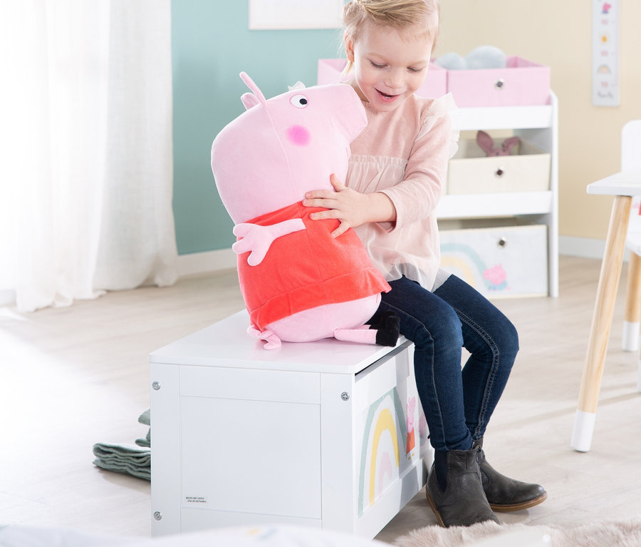 Mädchen sitzt auf einer weißen Spielzeugkiste und umarmt ein Peppa Pig Plüschtier.