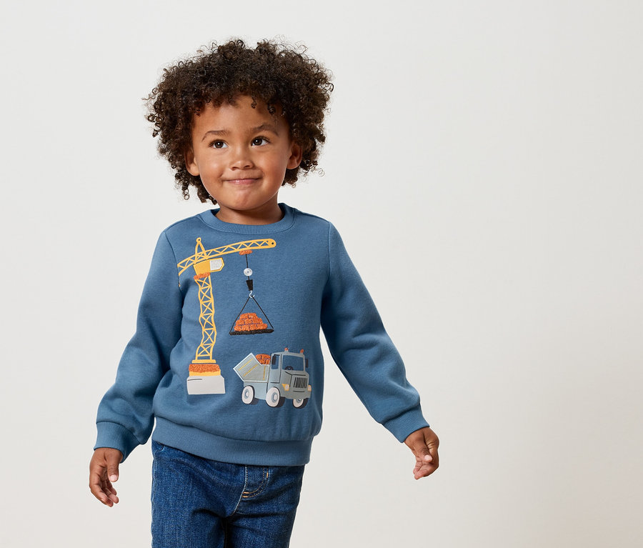 Ein lockiger Junge trägt ein blaues Sweatshirt mit einer gelben Kran- und Lastwagen-Applikation.