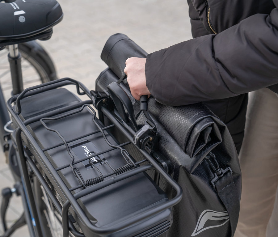 Eine Person befestigt eine schwarze FISCHER Gepäckträgertasche PLUS+ Terra an einem Fahrradgepäckträger.