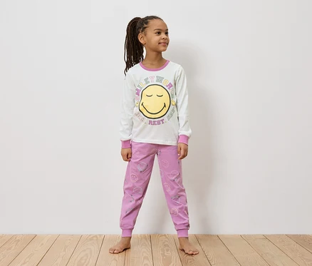 Mädchen mit Zöpfen steht barfuß in einem weißen, langärmeligen Smiley-Shirt und einer rosa Hose mit Smiley-Gesichtern.