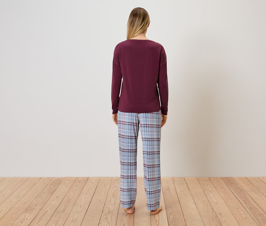 Eine Frau steht mit dem Rücken zur Kamera und trägt einen bordeauxroten Pullover und eine blau-karierte Pyjamahose.