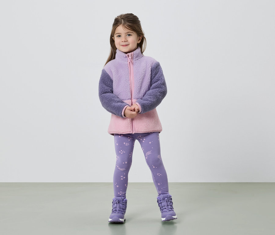 Ein Mädchen steht mit verschränkten Händen da und trägt eine Fleecejacke in Lila und Rosa, Leggings mit Sternen und lila Stiefel.