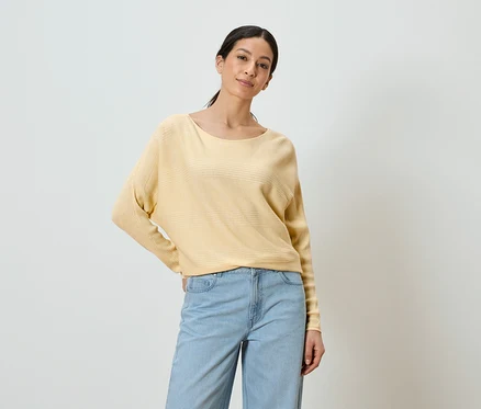Frau mit gelbem Pullover und blauen Jeans posiert vor einem weißen Hintergrund.