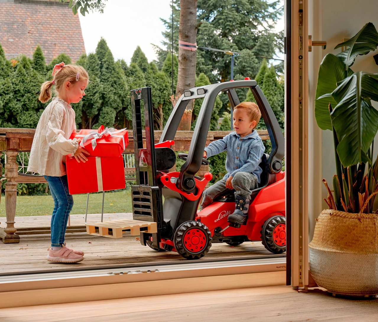 Mädchen trägt rotes Geschenk zum BIG Tretfahrzeug »Linde Gabelstapler«, auf dem ein Junge sitzt.