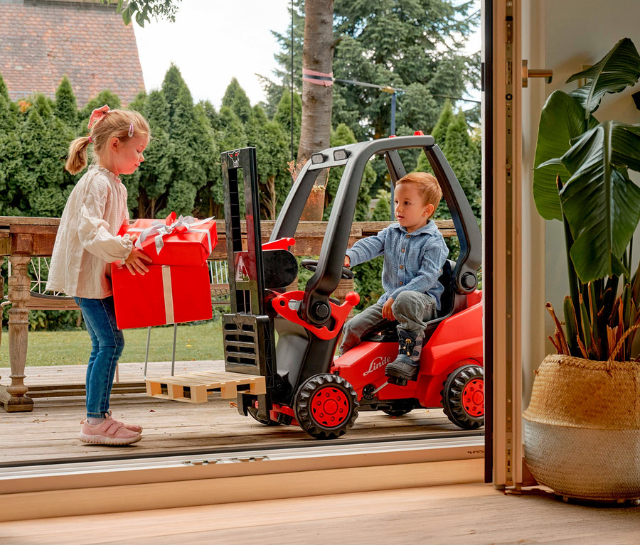 Mädchen trägt rotes Geschenk zum BIG Tretfahrzeug »Linde Gabelstapler«, auf dem ein Junge sitzt.
