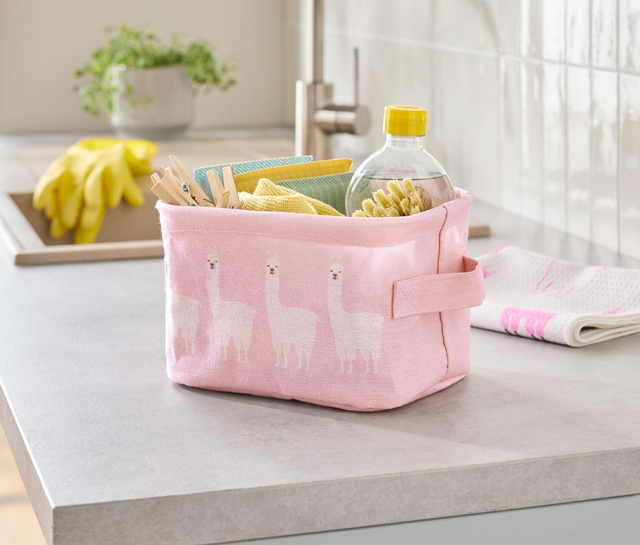 Auf einer Küchentheke steht eine rosa Stoffbox mit Lama-Muster, gefüllt mit Reinigungstüchern, Wäscheklammern und einer Flasche Reiniger.