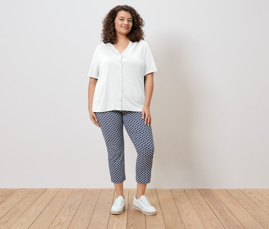 Frau mit lockigem Haar trägt eine weiße Hemdbluse und blaue gemusterte Hose, dazu weiße Schuhe.