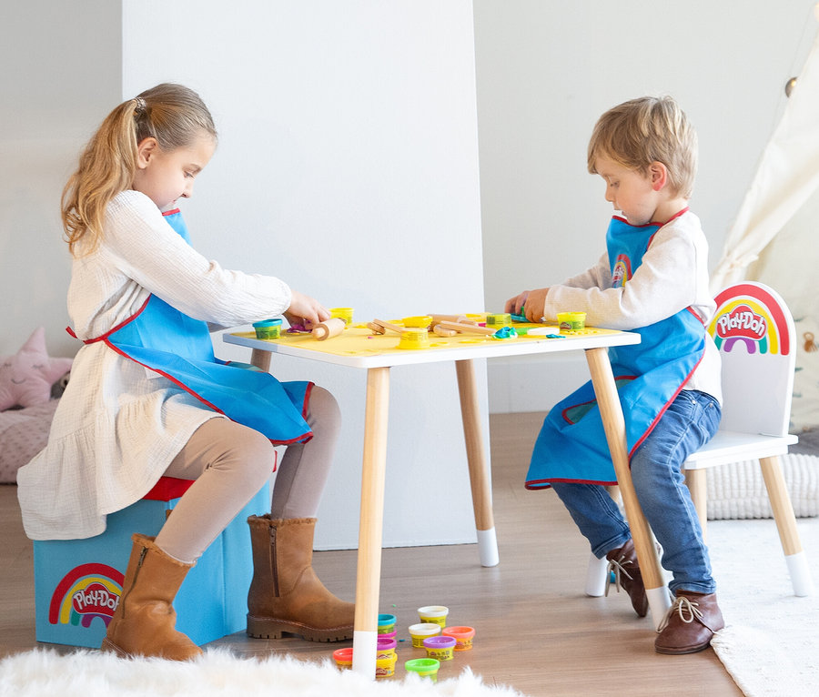 Zwei Kinder mit blauen Schürzen spielen an einem gelben Tisch mit Play-Doh. Ein Kind sitzt auf einer blauen Play-Doh-Spielzeugkiste.