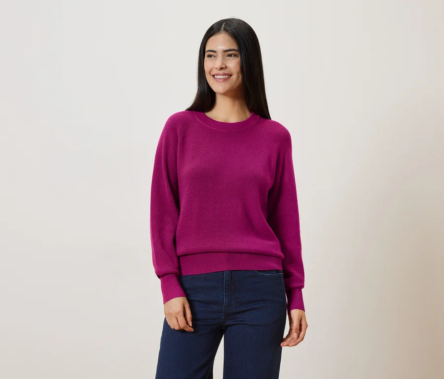 Frau mit dunklen, langen Haaren trägt einen pinken Pullover und blaue Jeans vor einem hellen Hintergrund.