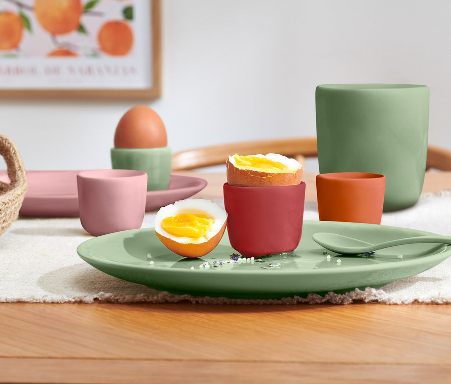 Ein Frühstücksarrangement auf einem Holztisch mit weißem Platzset zeigt verschiedenes farbenfrohes Geschirr. Ein weichgekochtes Ei in einem roten Eierbecher und ein halbiertes weichgekochtes Ei liegen auf einem hellgrünen Teller mit passendem Löffel. Weitere Gegenstände sind ein ganzes Ei in einem grünen Eierbecher, eine rosa Tasse und ein rechteckiger Teller, eine orangefarbene Tasse, eine große grüne Tasse und ein kleiner geflochtener Korb mit Eiern. Im Hintergrund hängt ein gerahmtes Bild mit Orangen.